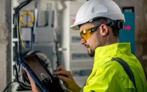 Man an electrical technician working in a switchboard with fuses, uses a tablet.